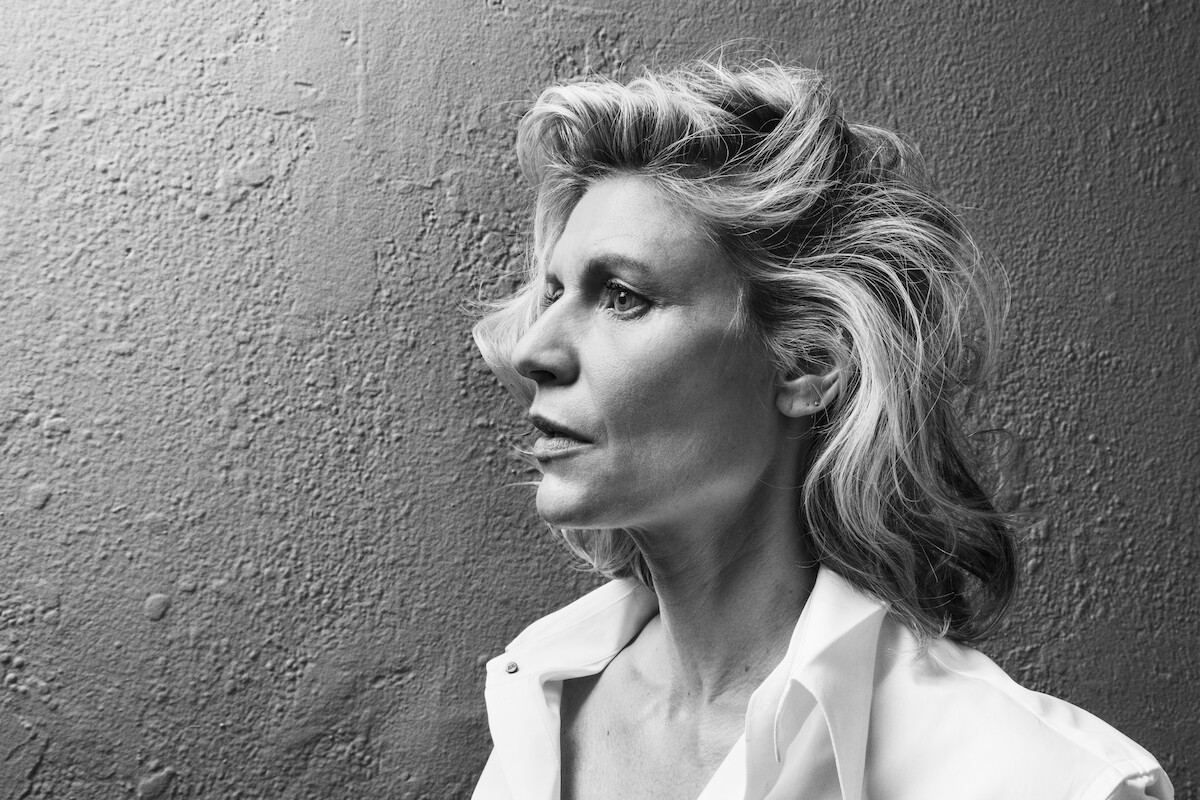 Black and white portrait of a woman with wavy hair in an open-collar shirt, gazing thoughtfully to the side against a textured wall, evoking a contemplative and classic mood.