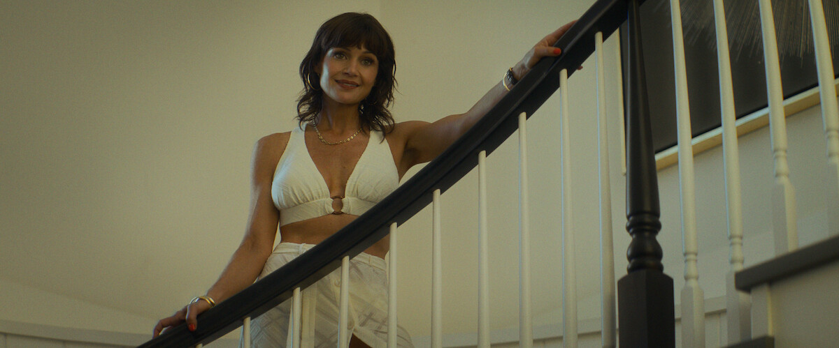 A woman in a white outfit stands on a curved staircase indoors, smiling, with one hand on the railing. The setting has neutral walls and framed art, giving a modern and welcoming home atmosphere.
