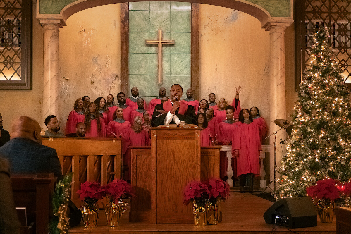 A gospel choir in red robes sings in a church decorated for Christmas with poinsettias and a lit tree. A preacher stands at the pulpit in front of a cross, addressing the congregation. Warm, festive atmosphere.