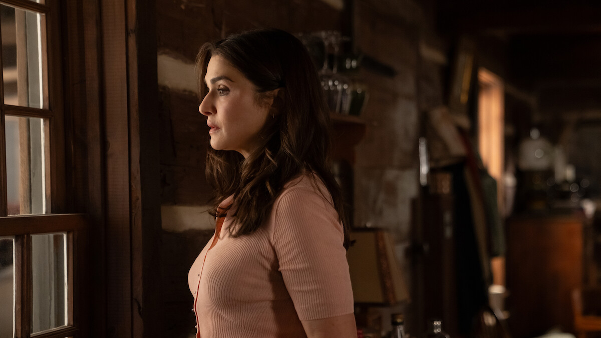 Woman with long brown hair stands indoors in a cozy wooden cabin, looking thoughtfully out the window. Warm lighting and rustic decor fill the background, creating a serene and intimate atmosphere.