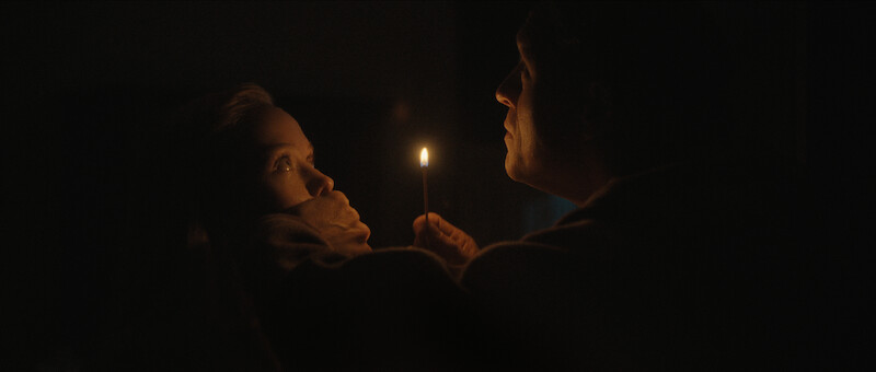 In a dark setting, a man holds a lit match close to his face while covering a woman's mouth, both illuminated by the small flame.