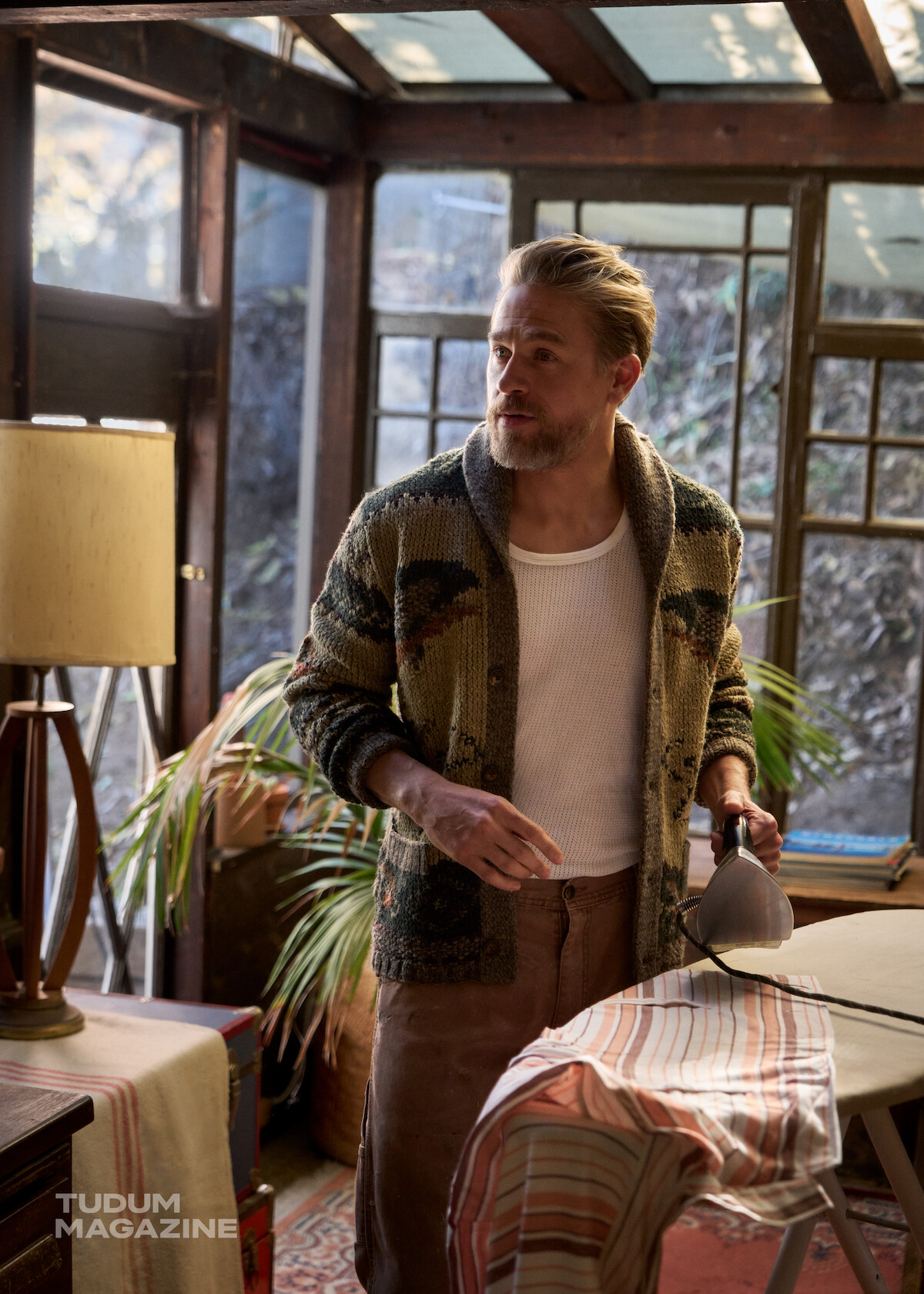 Charlie Hunnam stands in a sun room ironing a vintage shirt.