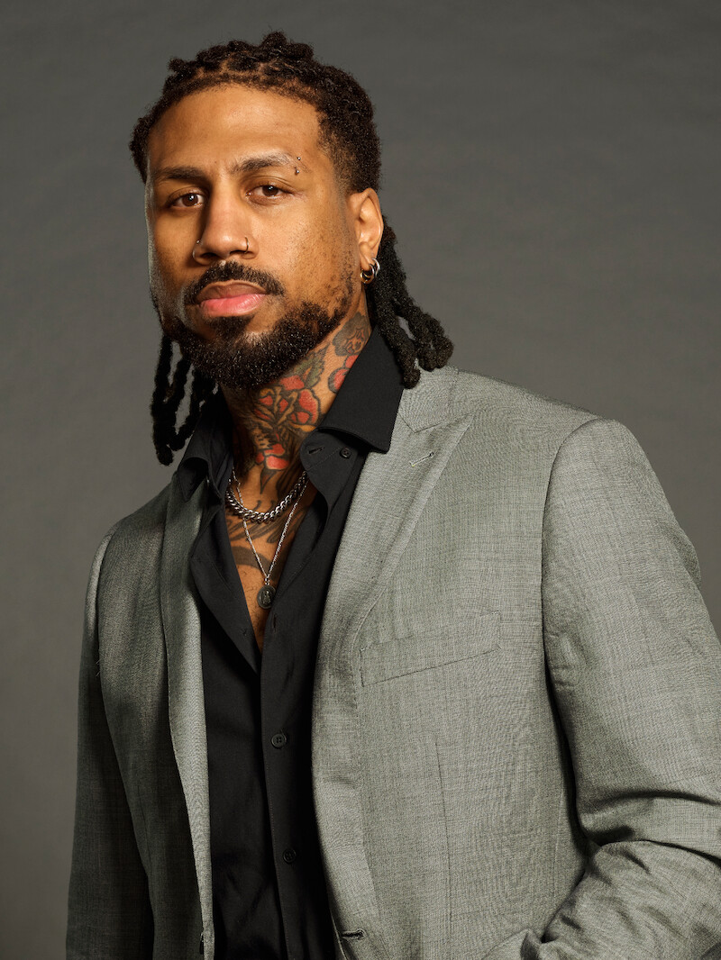 Homem com dreadlocks, barba e tatuagens no pescoço usando um terno cinza-claro e camisa preta, em frente a um plano de fundo neutro com iluminação suave, posa de forma confiante.