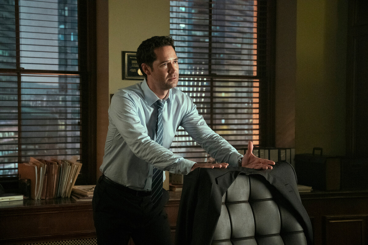 A man stands over a desk chair with his suit jacket draped on it. He is gesturing to someone out of frame.