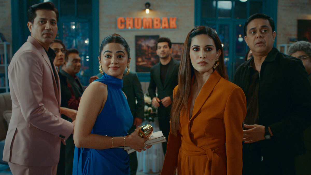 A group of well-dressed people at an indoor gathering or party, looking toward the camera with expressions of surprise or curiosity; modern decor and a sign reading "CHUMBAK" in the background.