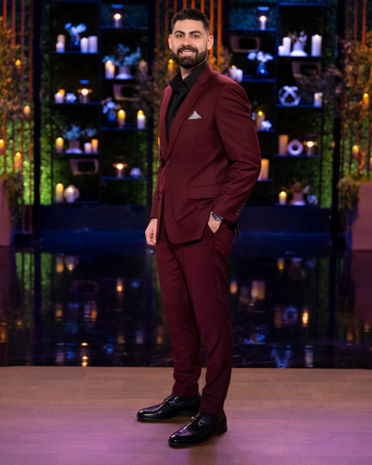Man with a beard in a burgundy suit stands on a polished stage with a dark, elegant backdrop filled with candles, shelves, and decorative plants, creating a formal and stylish atmosphere.