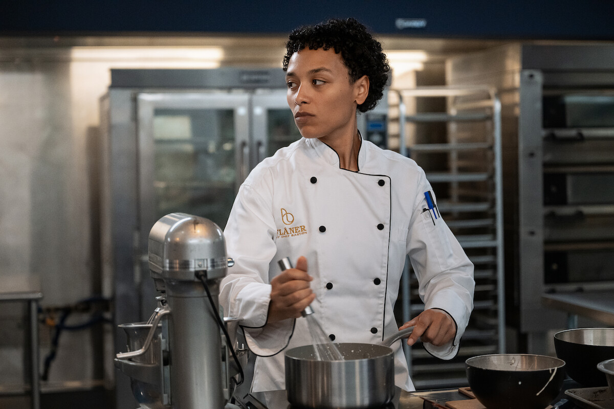 Chef in a white uniform mixing ingredients in a commercial kitchen, surrounded by baking equipment, stainless steel bowls, and industrial ovens in the background.