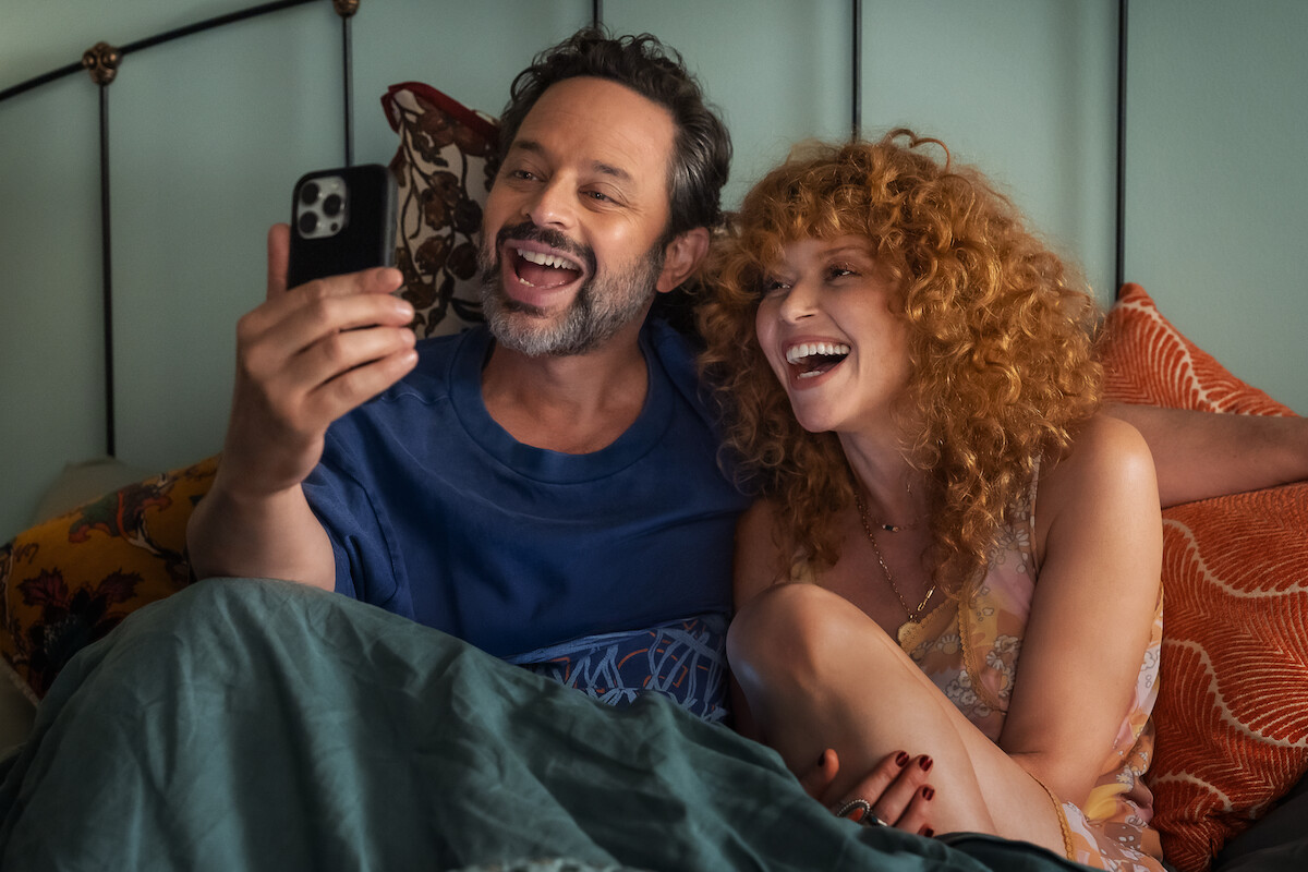 A man and woman sit together in bed smiling and laughing while taking a selfie or video call with a smartphone, creating a cozy and joyful atmosphere in a bedroom setting.