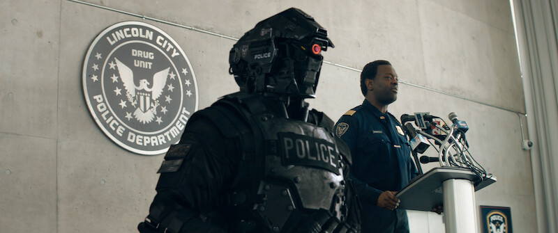 A policeman full body armor stands at a stage during a press conference in 'Code 8'