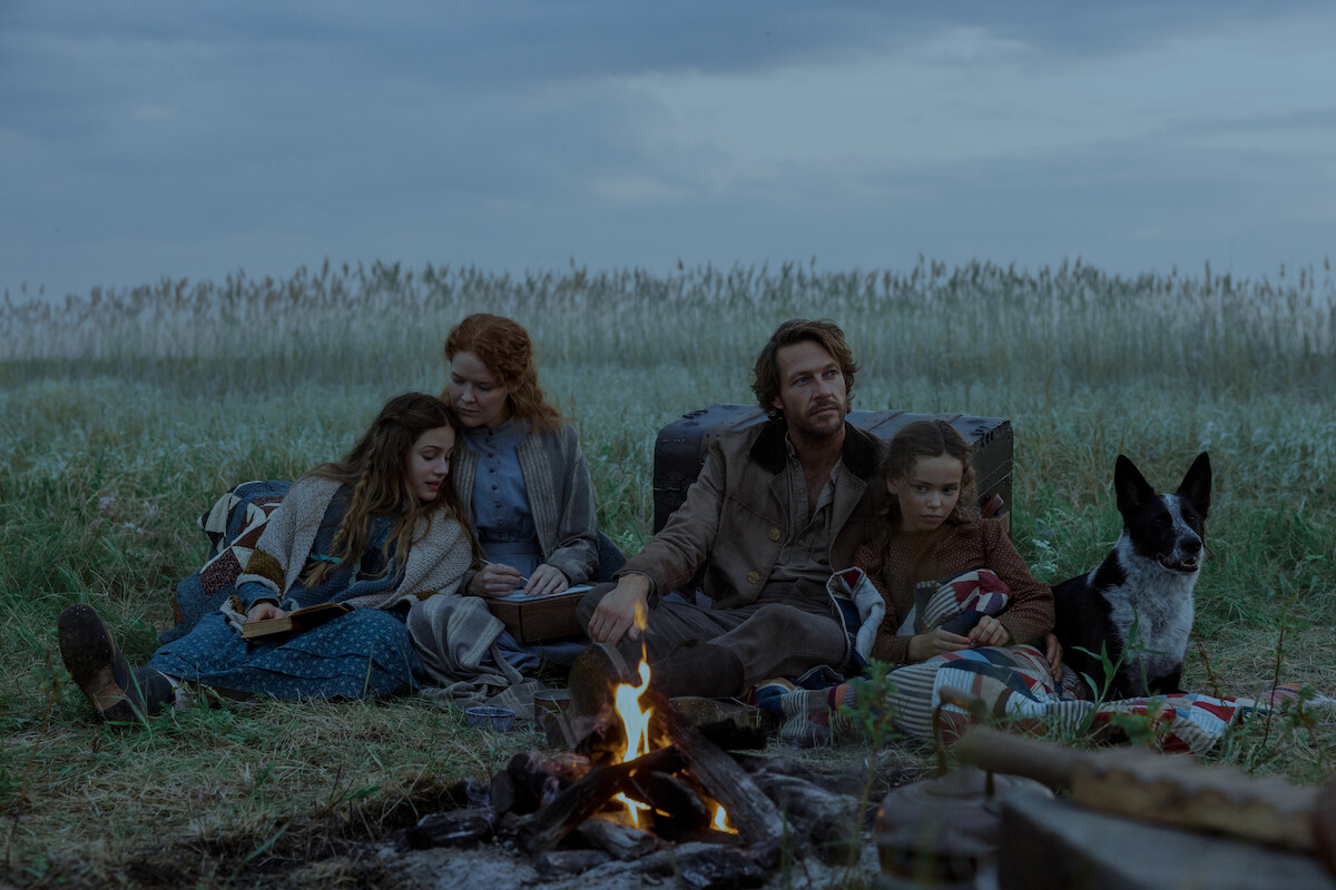 A family of four and a dog sit together by a campfire in a grassy field under a cloudy sky, surrounded by camping gear, suggesting a rustic outdoor setting and a calm, intimate atmosphere.