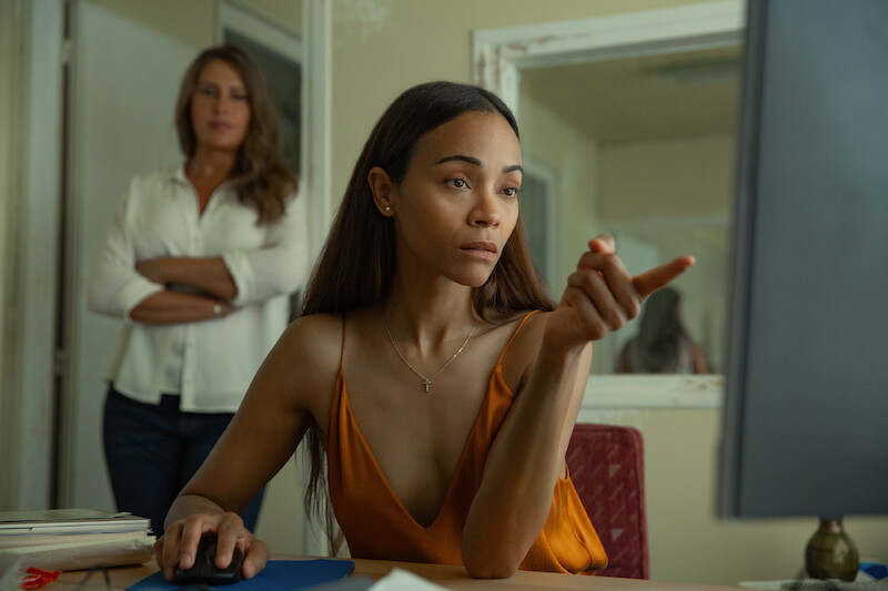 Emilia Pérez (Karla Sofía Gascón) stands behind Rita (Zoe Saldaña) in a white room.