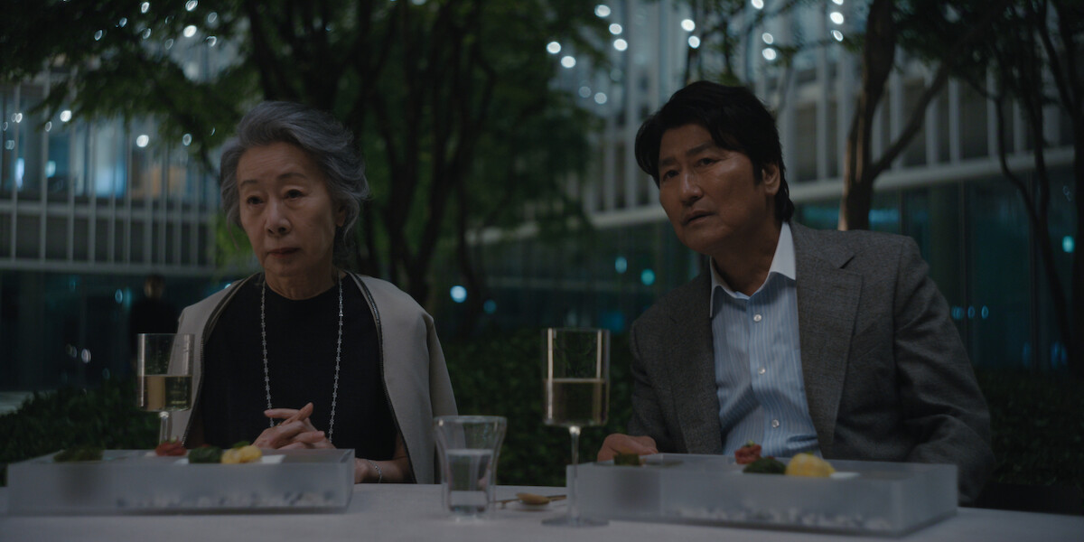 An elderly woman and a middle-aged man sit at an outdoor table in a modern, tree-filled courtyard at night, with glasses of wine, water, and plated food in front of them, surrounded by softly lit buildings.