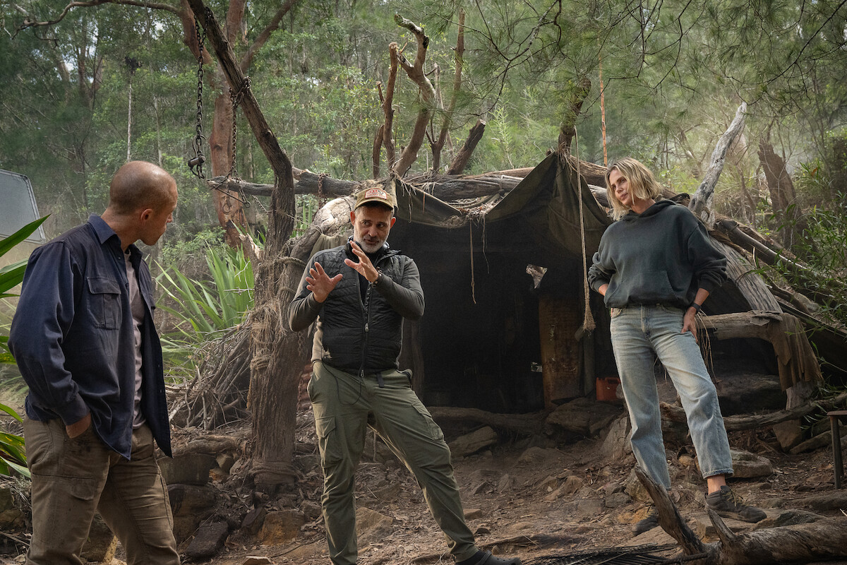 Three people stand and talk in a wooded outdoor setting, in front of a rustic makeshift shelter made of branches and tarps, surrounded by trees and natural vegetation.