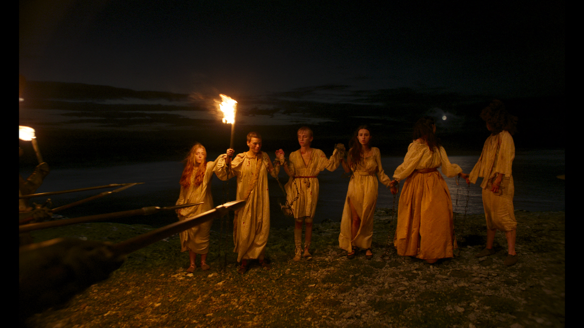 Six people in old-fashioned clothes stand in a line at night near water, holding hands, illuminated by torches while surrounded by armed figures pointing spears at them, creating a tense, dramatic scene.