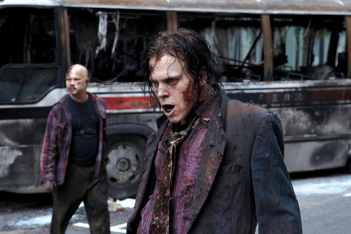 Two zombies in tattered clothing stand on a street in front of a burned, abandoned bus, with debris and destruction suggesting a post-apocalyptic urban environment.