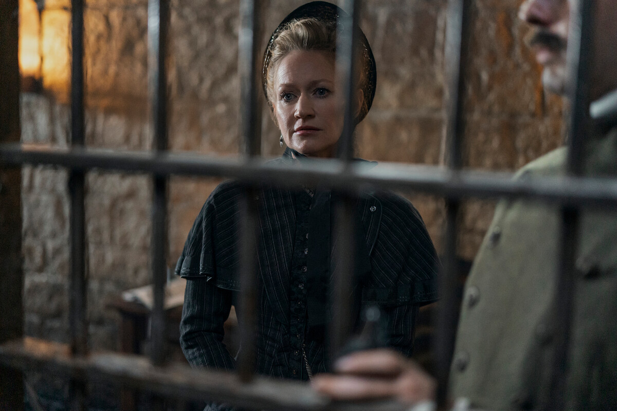 A woman in old-fashioned clothing stands behind iron bars in a dimly lit stone room, suggesting a historical prison scene with a somber and tense mood.