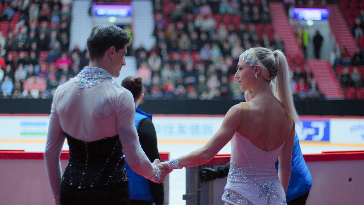 Two figure skaters in sparkly costumes hold hands near the rink barrier at a competition, preparing to perform in an indoor arena filled with spectators.