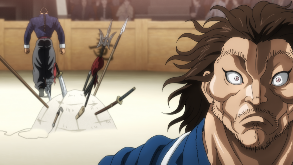 A shocked man with wild hair in a blue robe stands in an arena, with weapons impaled in the sand and a warrior walking away in the background. Spectators watch from behind a wooden barrier.