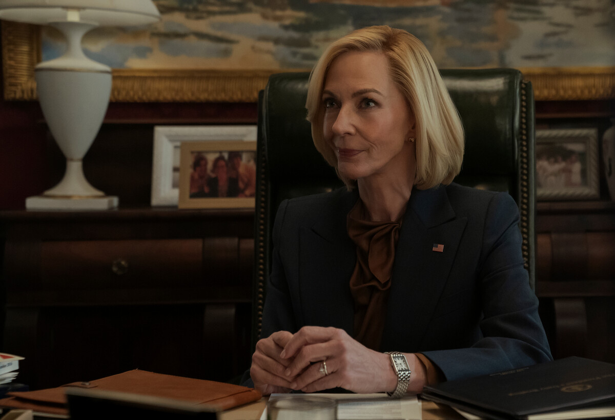 Woman in a suit sits at a desk in an office, hands clasped, with documents, family photos, a lamp, and a painting in the background, creating a professional and formal environment.
