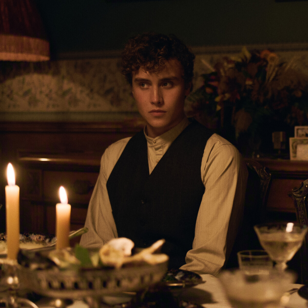 Joseph Zada as Cal Trask with curly hair wearing a vest sits at a candlelit dining table in a warmly lit, vintage room decorated with flowers and wallpaper, giving a thoughtful expression during an intimate dinner setting.