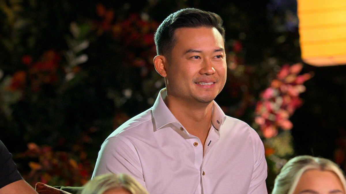 A man in a white shirt smiles while sitting outdoors at night, surrounded by blurred people and colorful foliage, creating a warm, relaxed, and cheerful atmosphere with soft lighting.