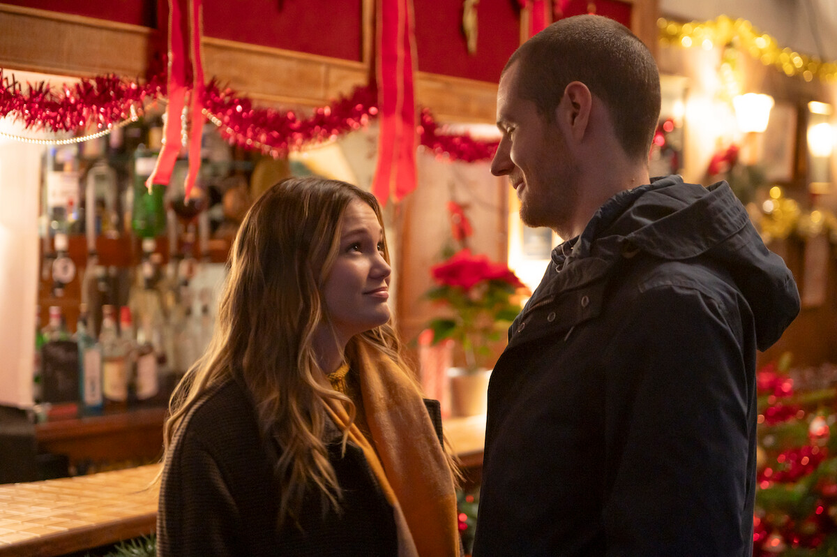 Olivia Holt as Sophia Martin and Connor Swindells as Nick O’Connor look at each other with Christmas decor behind them in an image from ‘Jingle Bell Heist.’