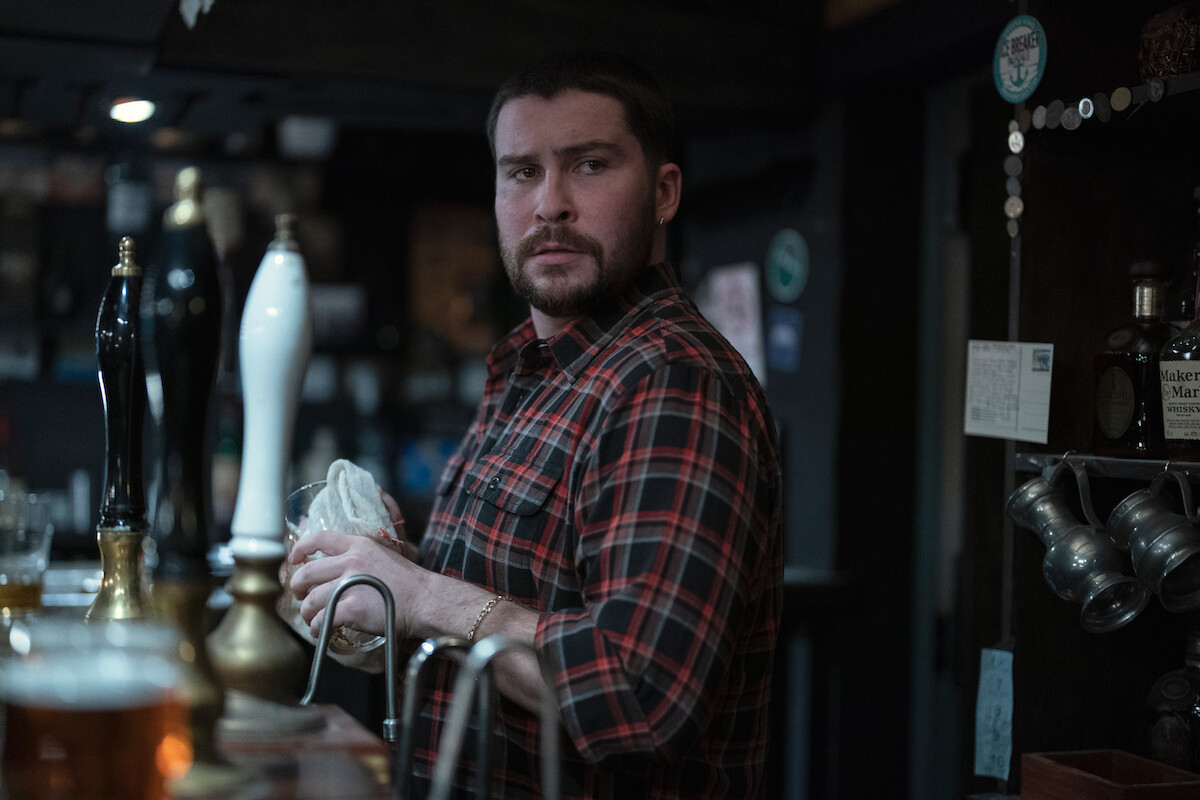 Daniel Portman as Stuart wears a plaid shirt and washes a dish behind a bar in Season 6 of ‘Black Mirror.’