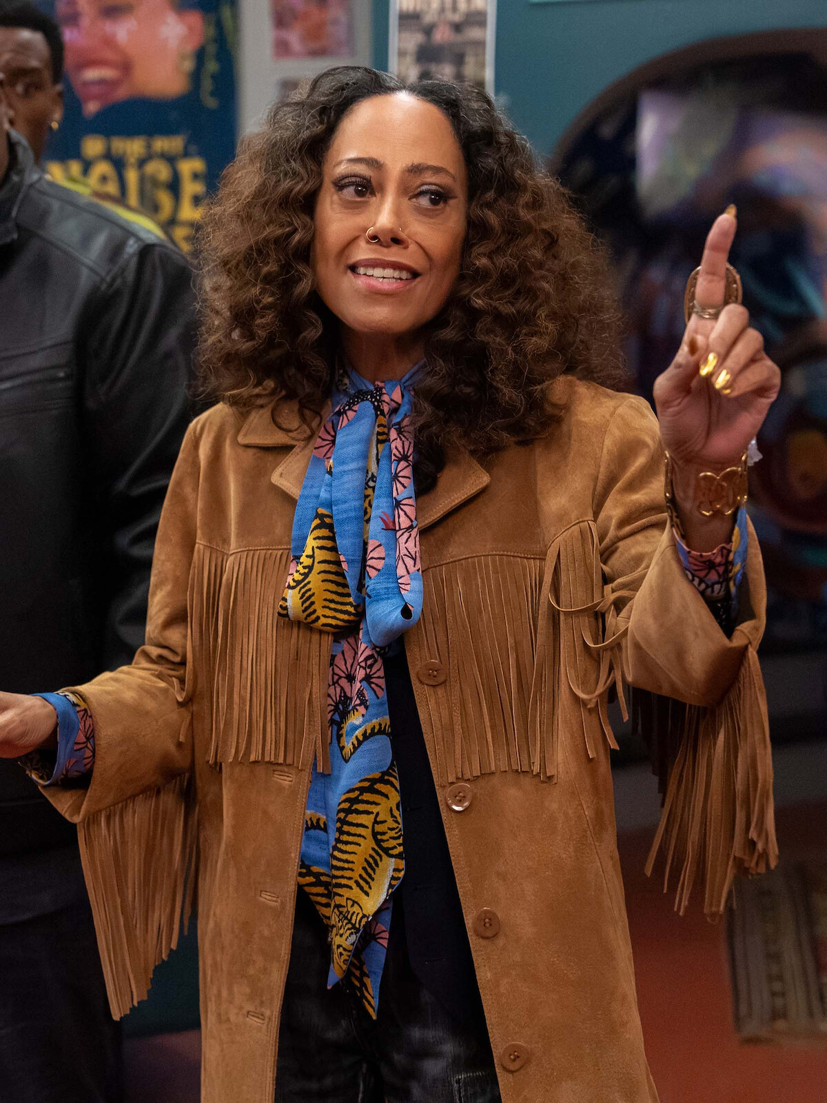 Woman with curly hair wearing a brown fringed jacket and colorful patterned scarf standing indoors, gesturing with both hands, with posters and people visible in the background.