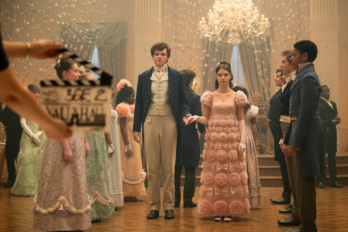 Cena de época com homens e mulheres elegantemente vestidos em um baile, sob um lustre ornamentado, em um grande salão. Em primeiro plano, a equipe de filmagem segura uma claquete para marcar a cena.