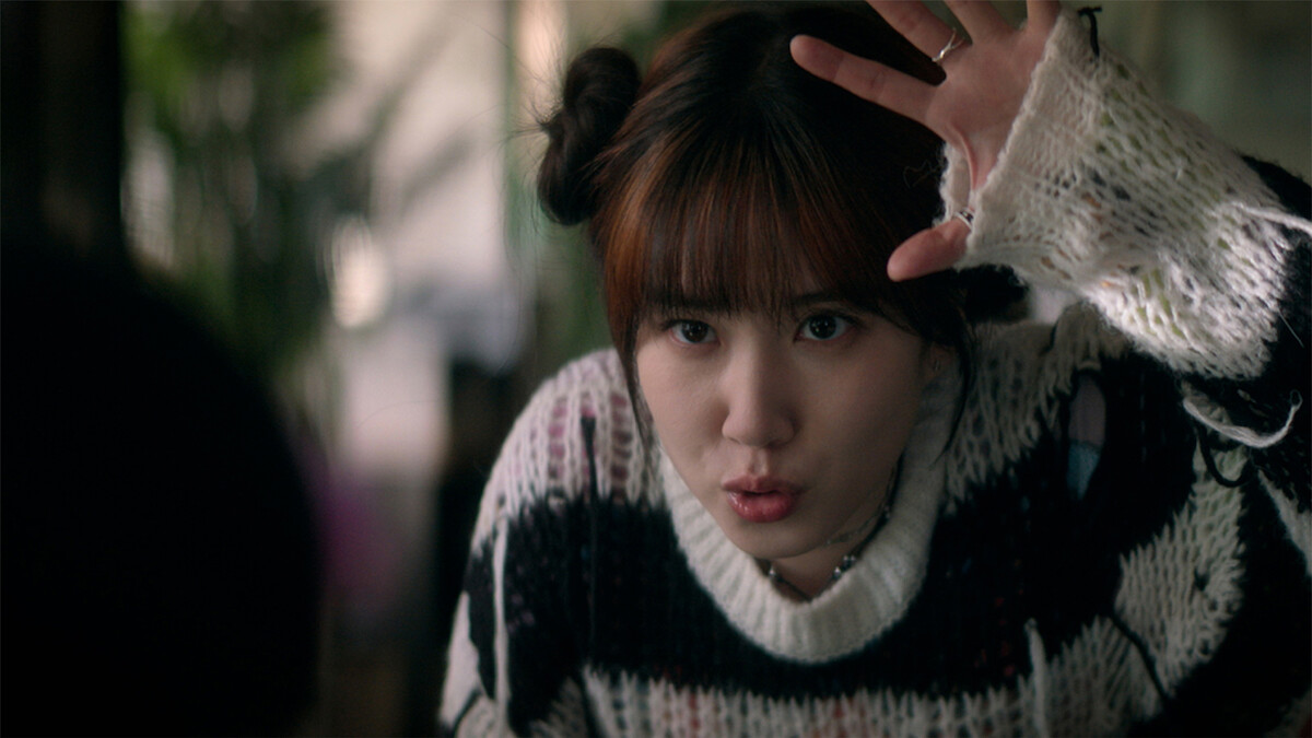 Young woman indoors with plants in the background, wearing a black and white sweater with frayed sleeves, hair in two buns, raising her hand and making a playful expression, as if acting or interacting animatedly.