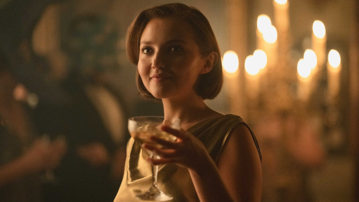 Woman in an elegant dress holding a champagne glass at a formal event, with soft lighting and chandeliers in the background, creating a warm, sophisticated atmosphere.