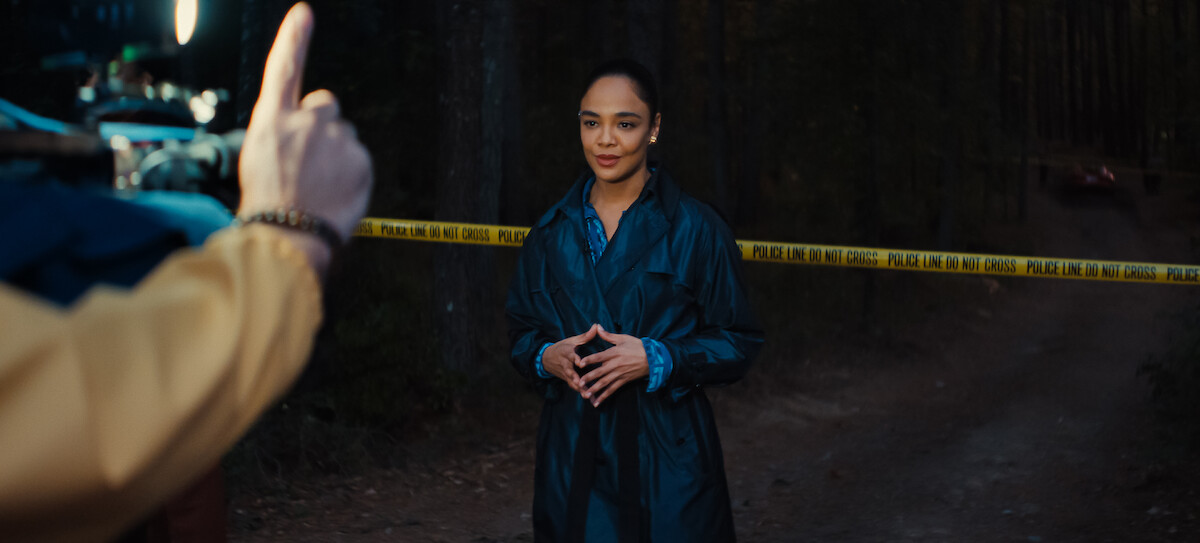 A news reporter stands in front of police tape in a dark forest while being filmed.