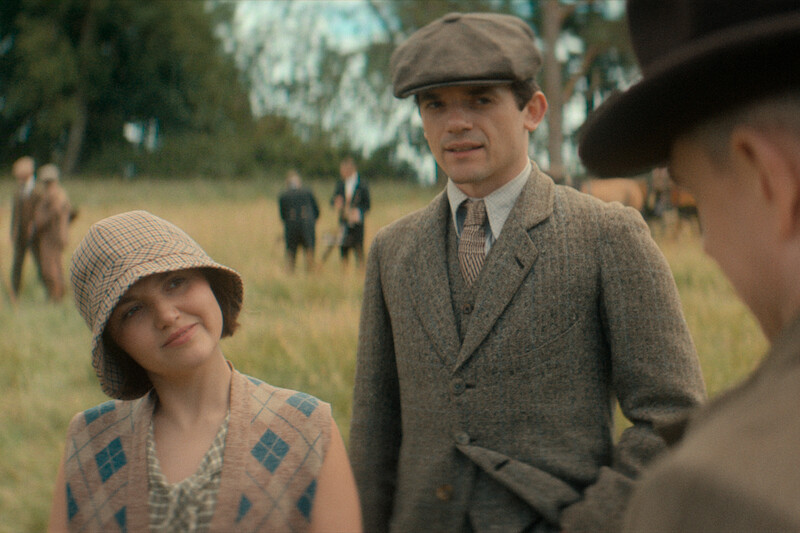 A man and woman in vintage clothing converse with another man outdoors in a grassy field; people and trees in the blurred background suggest a relaxed, early 20th-century countryside gathering. Mood is warm and nostalgic.