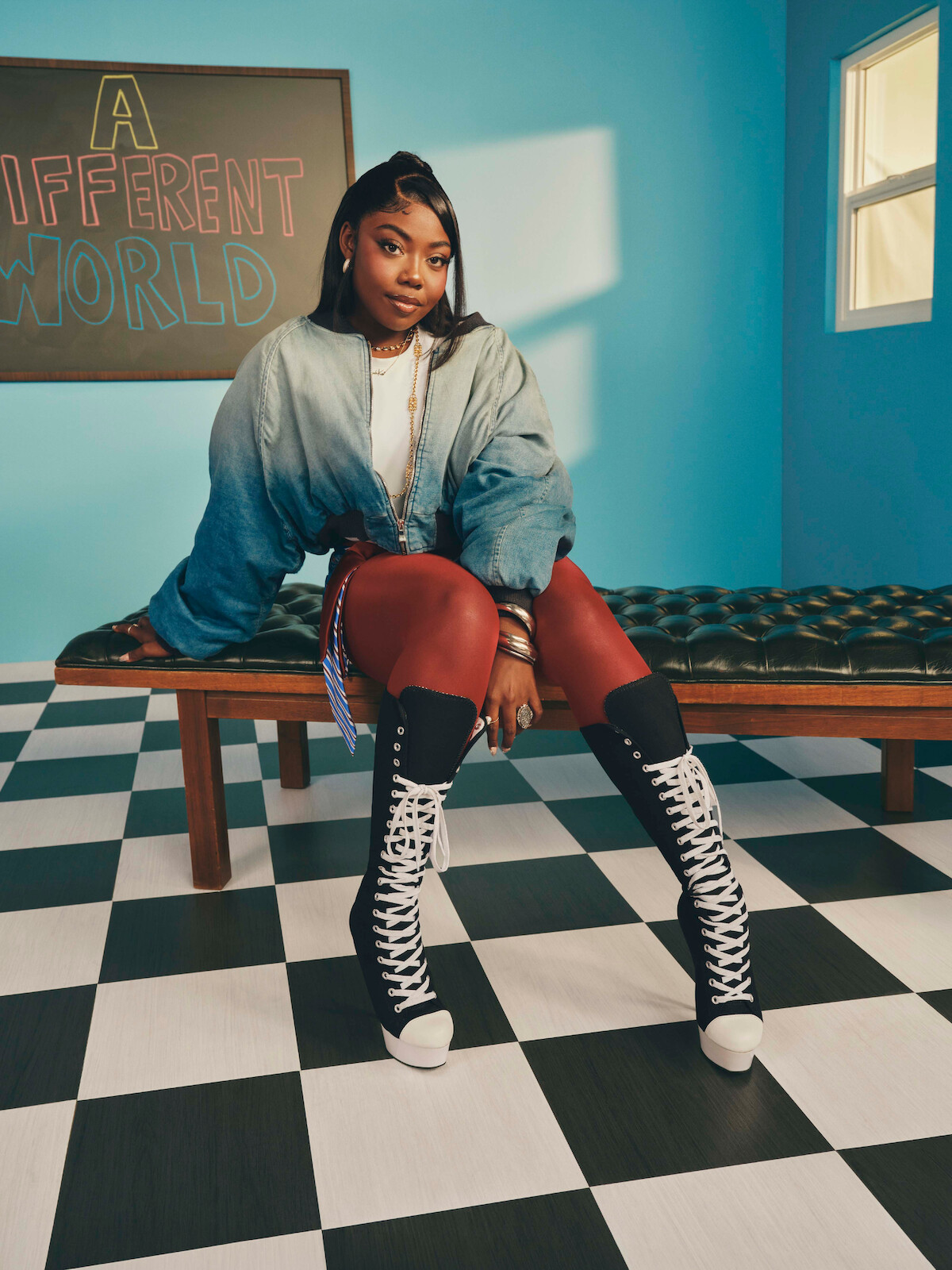 A woman sits on a bench in a retro-style room with checkered floor, blue walls, and a sign reading "A Different World." She wears a denim jacket, red leggings, and tall black-and-white lace-up boots.