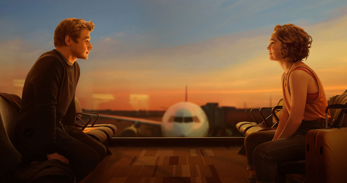 Ben Hardy and Haley Lu Richardson sit opposite one another on an airport terminal floor, with a plane between them outside the window.
