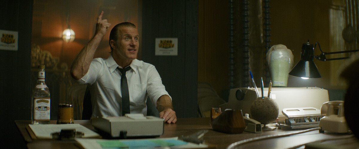 Man in a white shirt and tie sits at a cluttered office desk with papers, a drink, and a bottle, gesturing energetically. The office is dimly lit with vintage decor, a typewriter, desk lamp, and industrial pipes in the background.