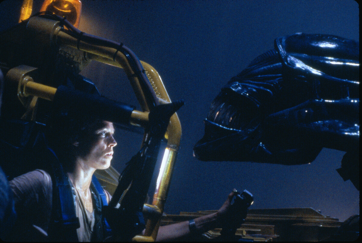 A person inside a yellow mechanical exosuit faces a large black alien creature in a dark, industrial, sci-fi environment with dramatic lighting.