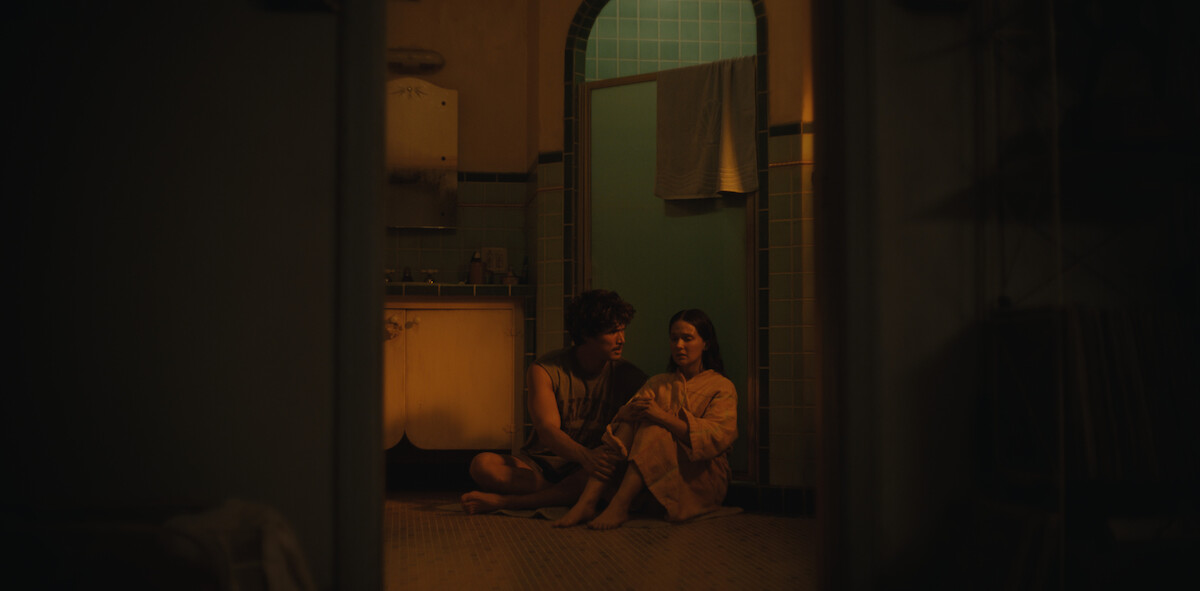 Two people sit close together on a bathroom floor at night, lit by warm, soft lighting. The scene appears intimate and quiet, with vintage-style fixtures and a towel hanging over a glass door in the background.