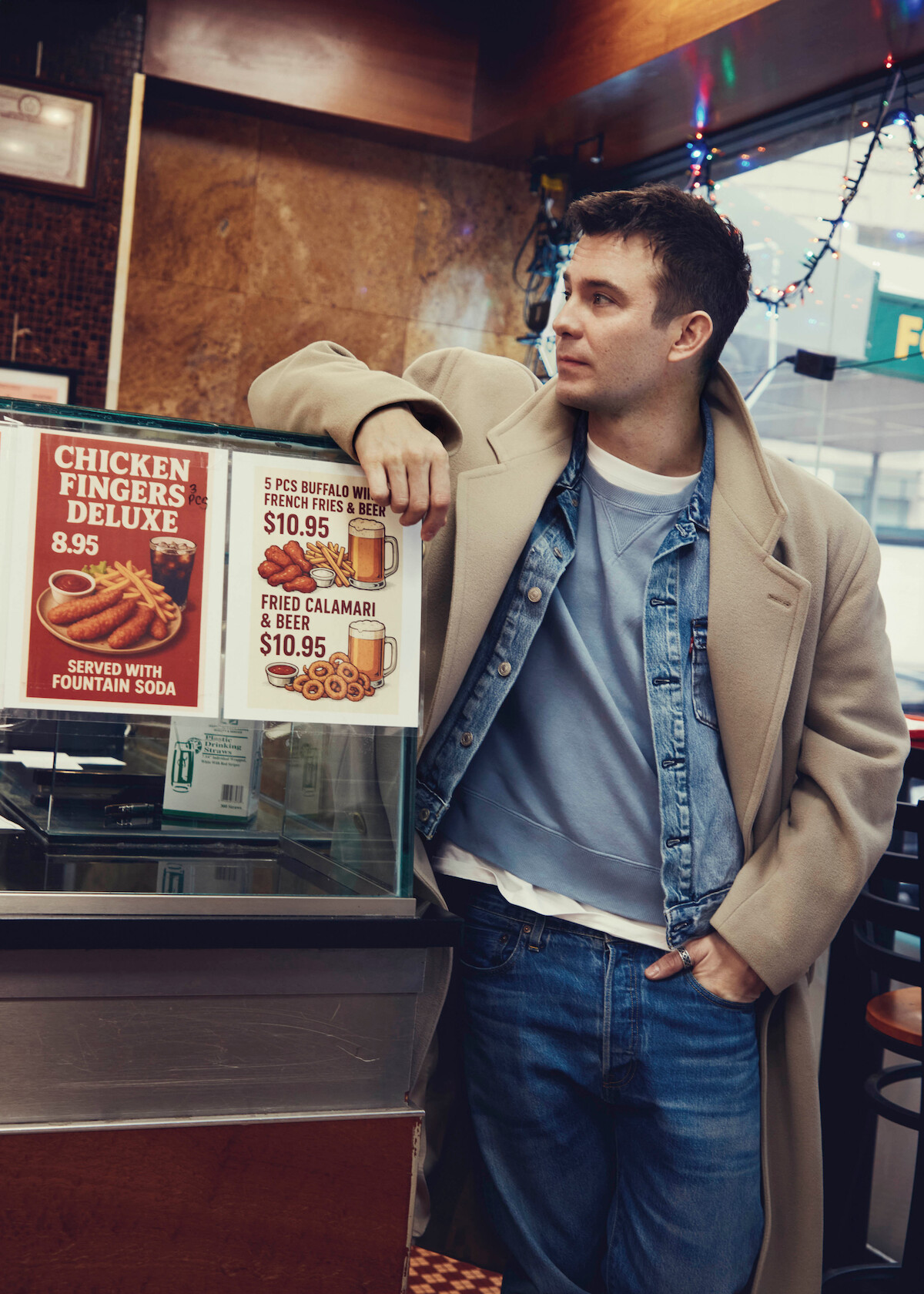 Um homem usando roupas casuais em tons de azul e branco, com um casaco bege, está dentro de uma lanchonete apoiado em um balcão próximo a placas de menu com opções como tiras de frango e lula. O ambiente tem estilo urbano, iluminação suave e luzes decorativas penduradas.