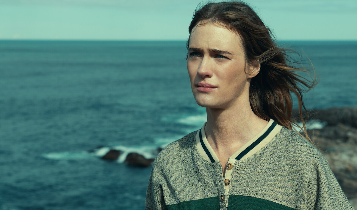 Person with long hair standing outdoors near the ocean, wearing a gray and green sweater. The sea and rocky shore are in the background under a clear sky, creating a serene coastal scene.