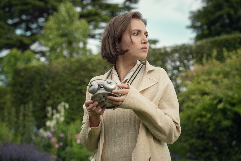 A woman in a beige coat stands in a lush garden holding a metal object, looking off to the side with a thoughtful expression. The scene feels mysterious and serene, surrounded by greenery and soft natural light.