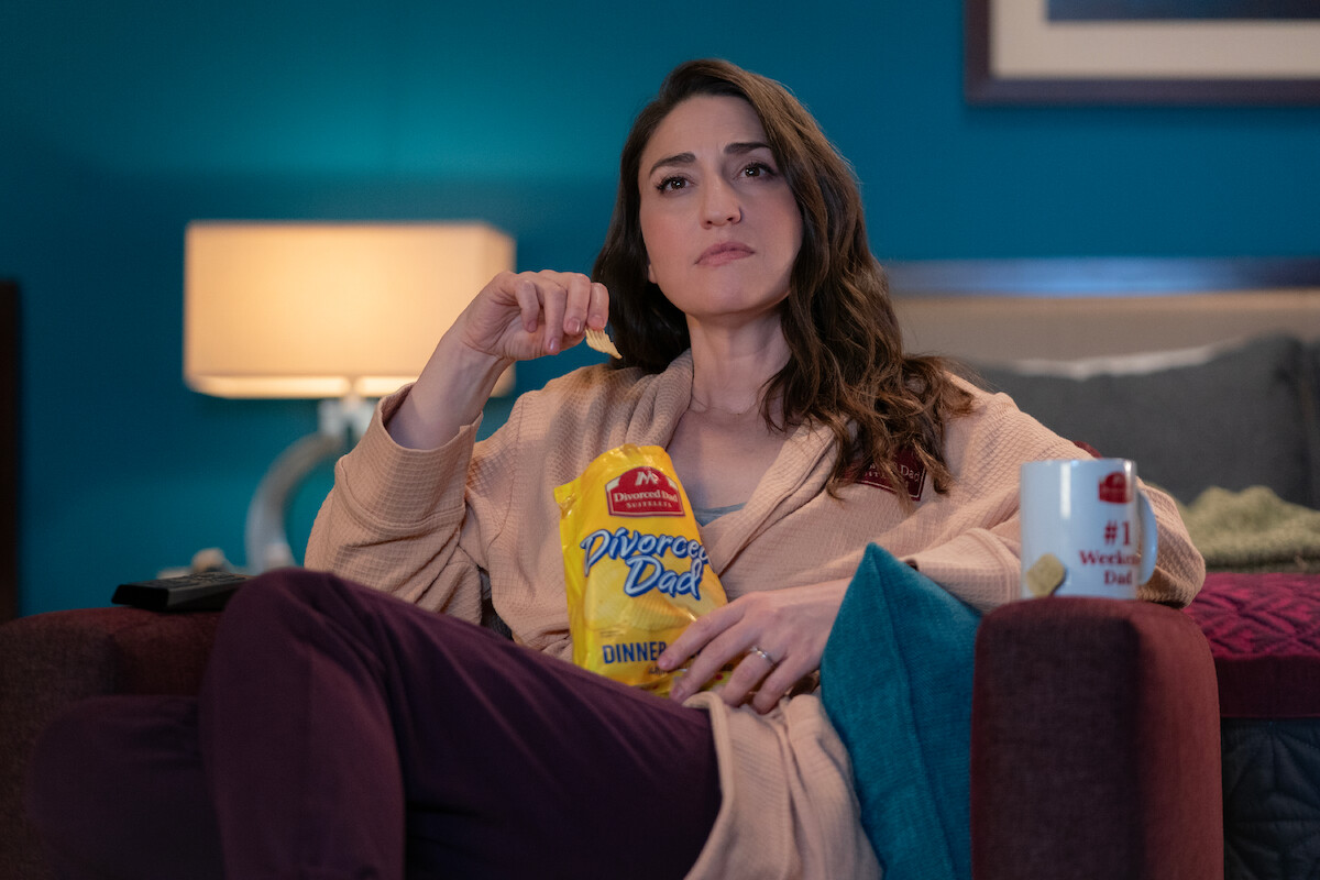 Sara Bareilles as Dawn wears a robe and eat snacks in a hotel room in season 3 of ‘Girls53va’