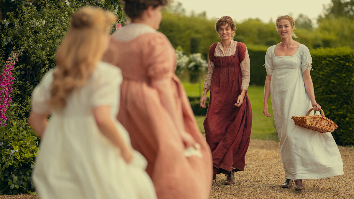 Four women in Regency-era dresses walk and smile outdoors on a garden path, surrounded by greenery and flowers, with one carrying a wicker basket.