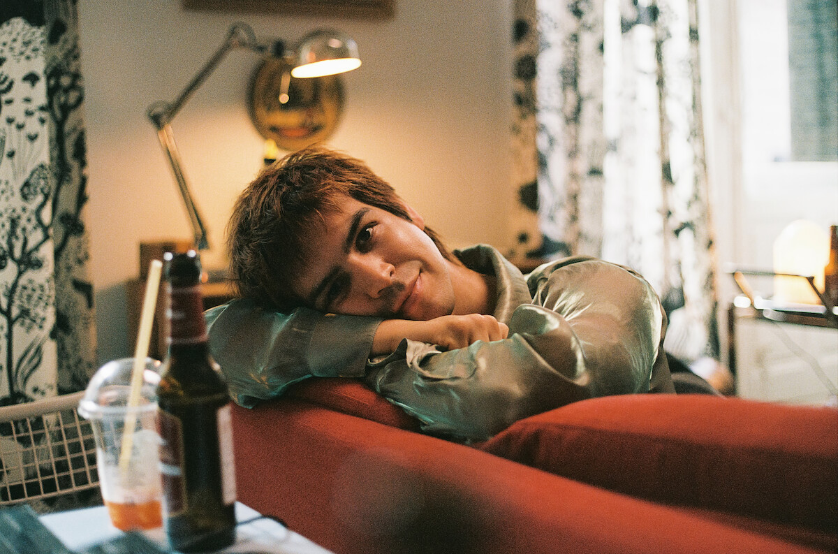 Person with short hair relaxing on a red couch in a cozy, warmly lit living room with patterned curtains, a table lamp, a drink cup, and a bottle in the foreground, creating a calm, intimate atmosphere.