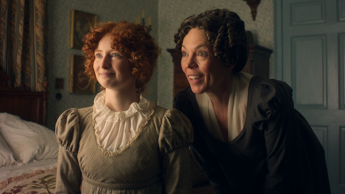 Two women in historical clothing stand indoors by a bed in a vintage-decorated bedroom, smiling as they look toward the window, with soft natural light illuminating their faces and period furniture visible in the background.
