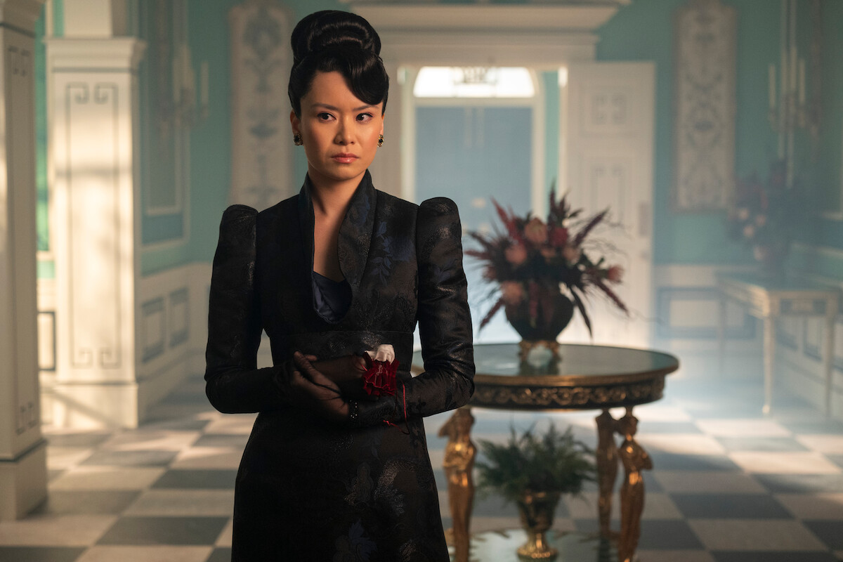 Woman in an ornate black gown stands with composed expression in an elegant, richly decorated room with checkered floor, teal walls, and a gold-trimmed table adorned with a floral arrangement in the background.