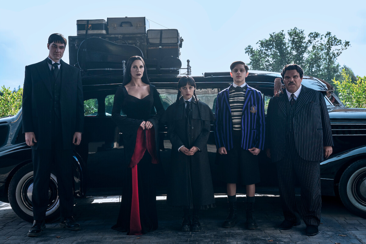 Joonas Suotamo as Lurch, Catherine Zeta-Jones as Morticia Addams, Jenna Ortega as Wednesday, Isaac Ordonez aș Pugsley Addams, Thing, and Luis Guzmán as Gomez Addams stand in front of their car in Season 2 of ‘Wednesday’