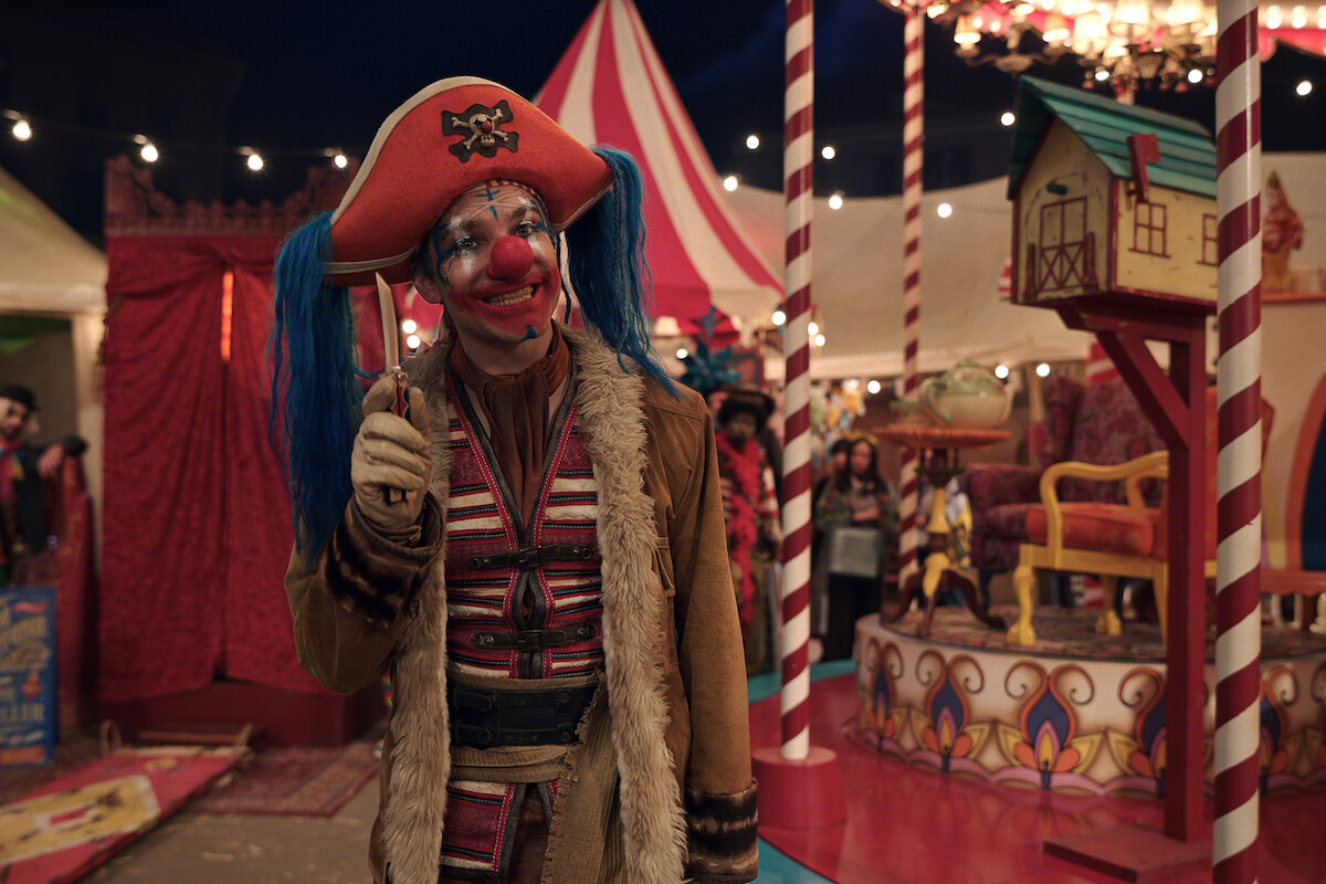 A clown in a red pirate hat, blue pigtails, and colorful costume stands smiling in a lively circus setting with striped tents, bright lights, festive decor, and people in the background.