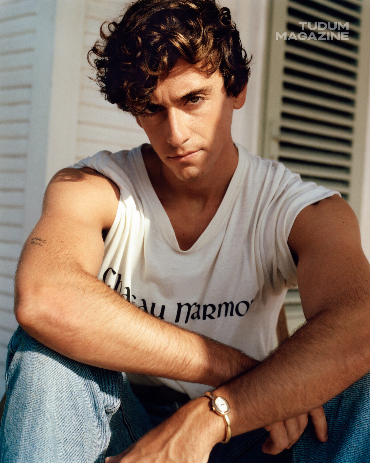 Young man with wavy brown hair sits casually in front of a white shuttered window, wearing a white t-shirt and jeans, looking intently at the camera in natural sunlight. "Tudum Magazine" text appears in the top corner.