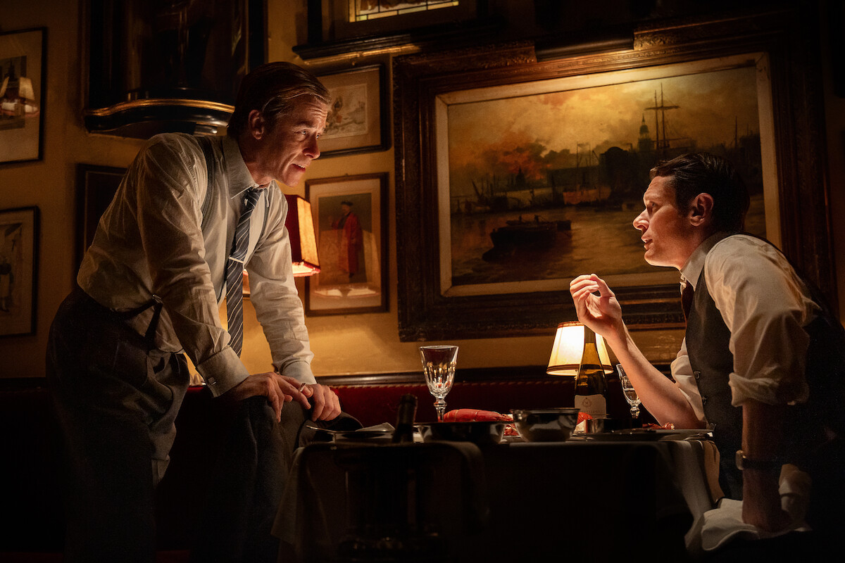 Two men in a dim, wood-panelled restaurant lean in and talk intently over a candlelit table.